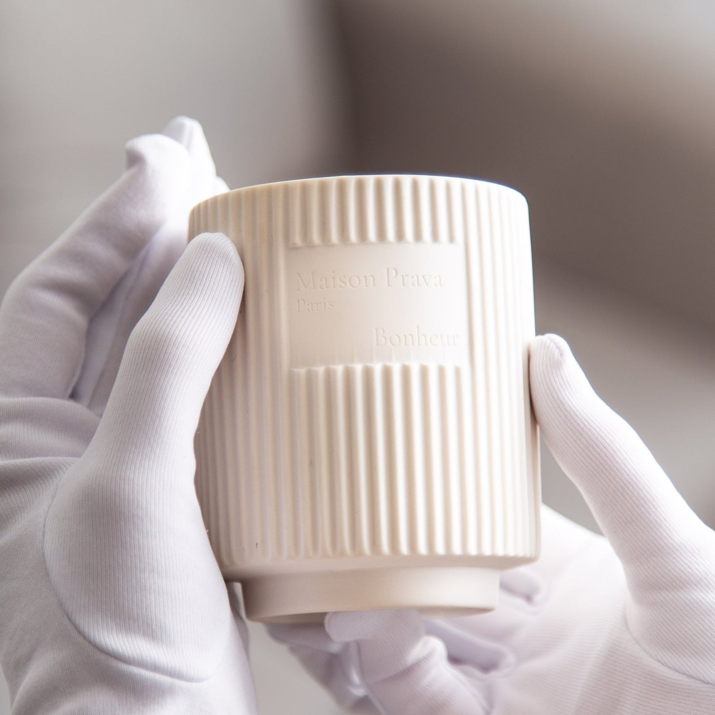 Beige textured ceramic luxury candle held by a person wearing white gloves against a neutral background