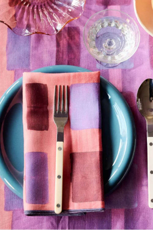 Plated setting with a checkered pink linen napkins, fork, and spoon on a purple checkered tablecloth.