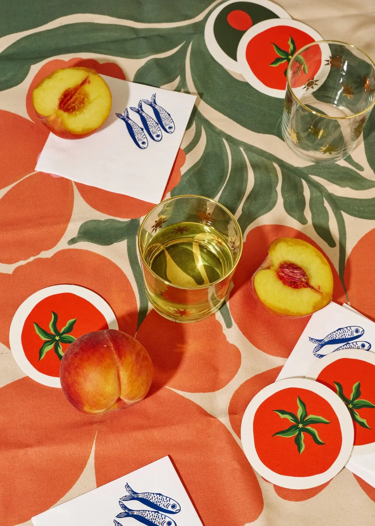 Table setting with peach slices, a glass of white wine, and fun tomato coasters on a decorative tablecloth.