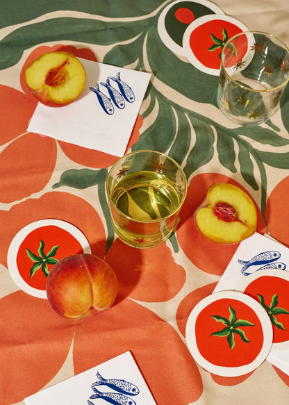 Table setting with peach slices, a glass of white wine, and fun tomato coasters on a decorative tablecloth.